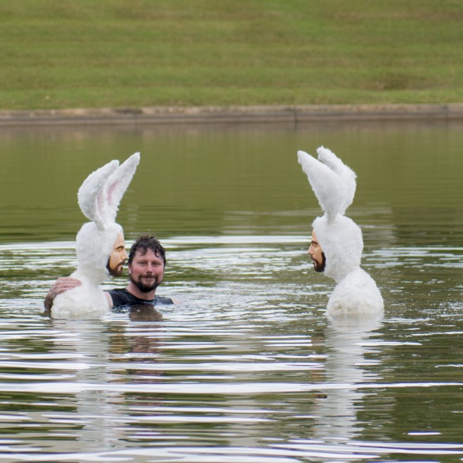 An artist swims in the lake with a water feature sculpture of two men in bunny suits