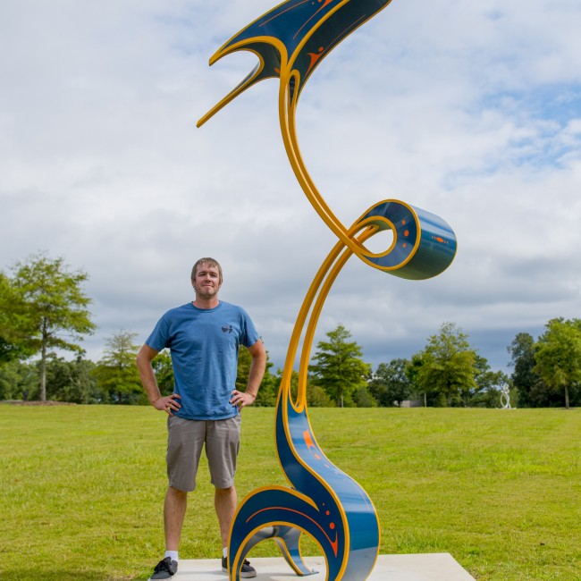 Artist Luke Achterberg stands next to his outdoor sculpture with painted curves