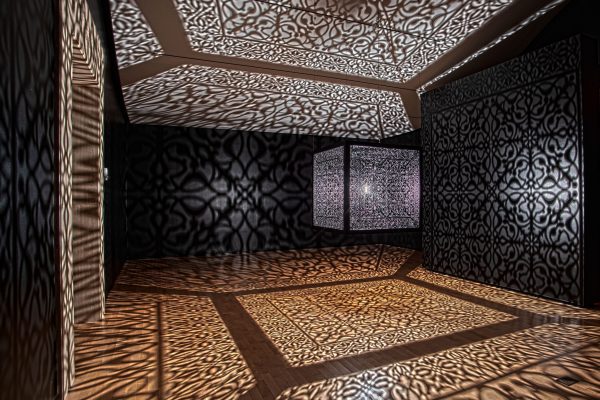 A large metal cube suspended from the ceiling, casting a cool complex pattern.