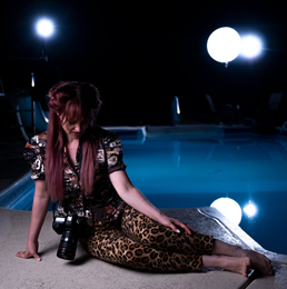 A woman sits in front of a pool staged for a night photoshoot, looking down at her camera.