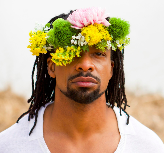 A man with dark braids and medium skin wears a flower crown.
