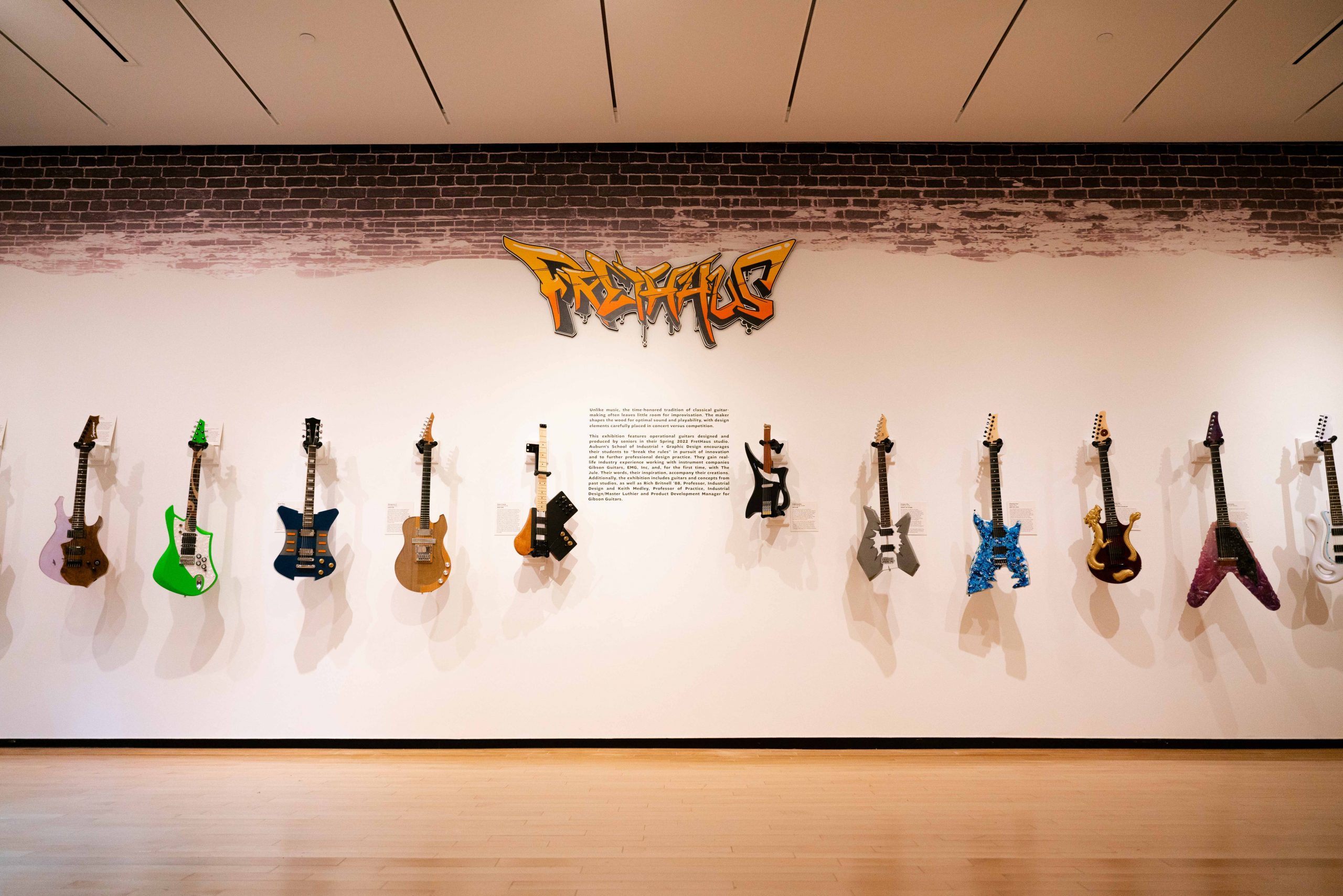 Uniquely decorated guitars hang on a wall with text describing the exhibition in the center and FretHaus in stylized lettering above