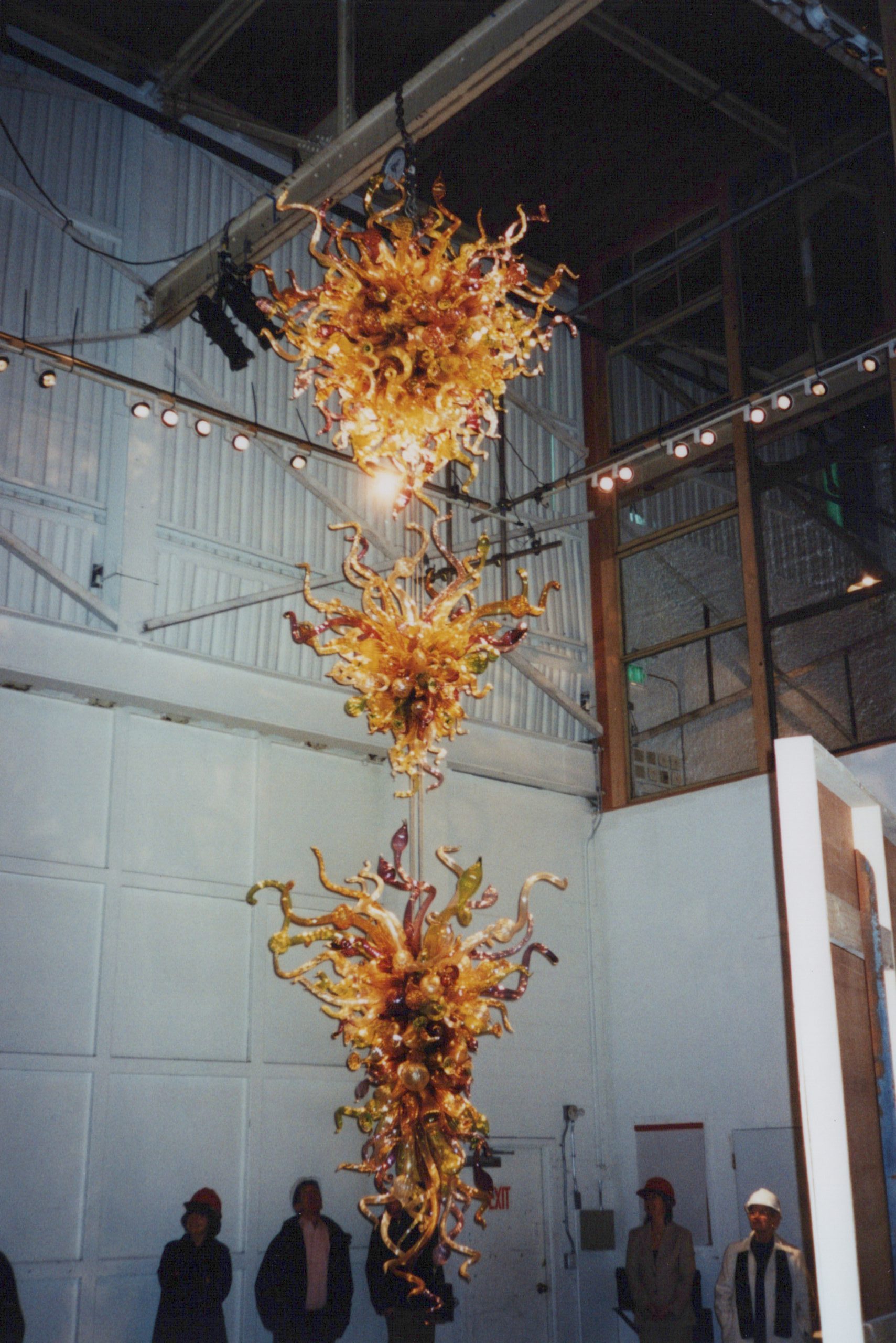 An intricate glass sculpture hangs in a warehouse while people below look at it.