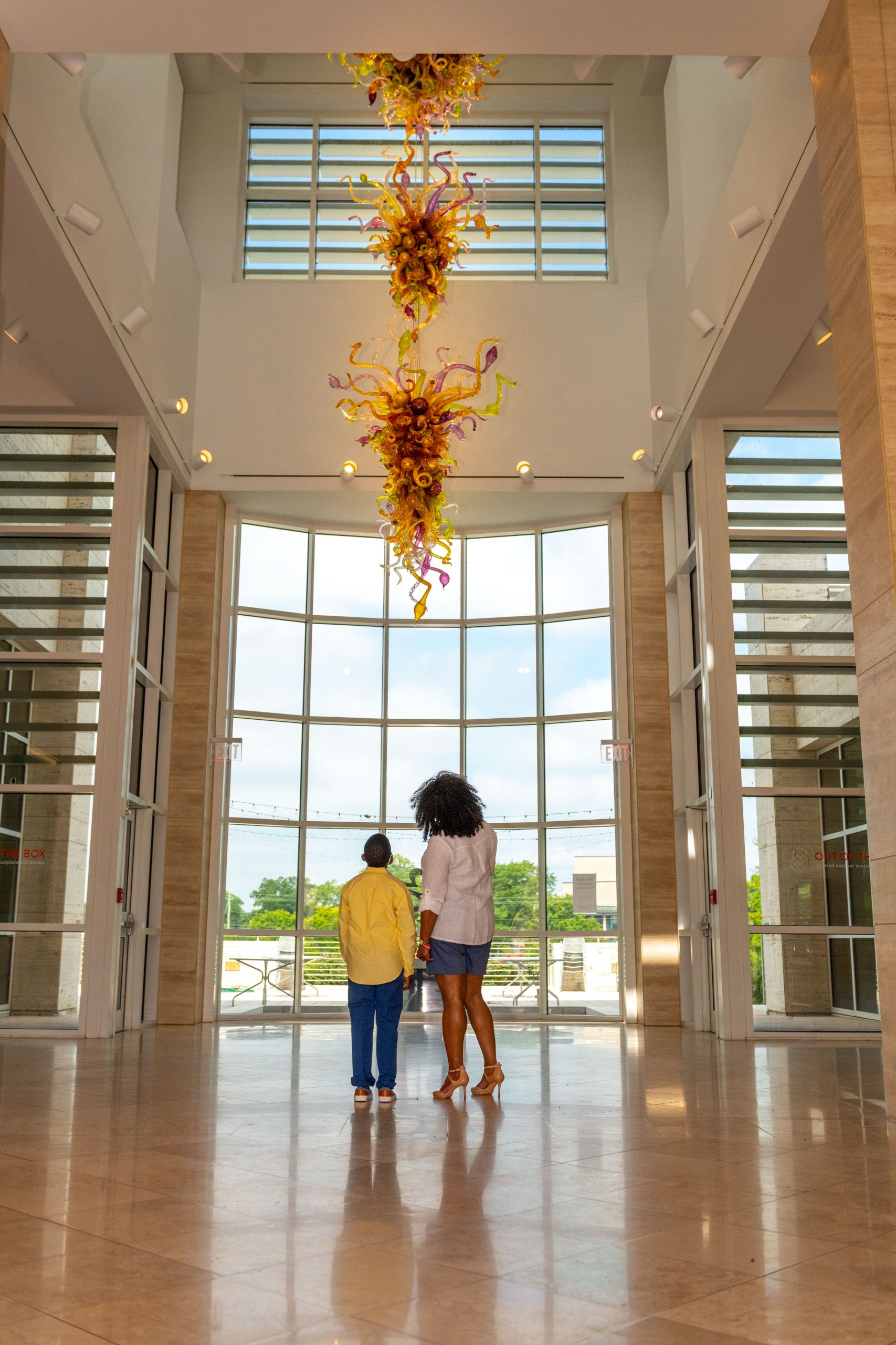 A woman and child look up at a glass sculpture that hangs from the ceiling.