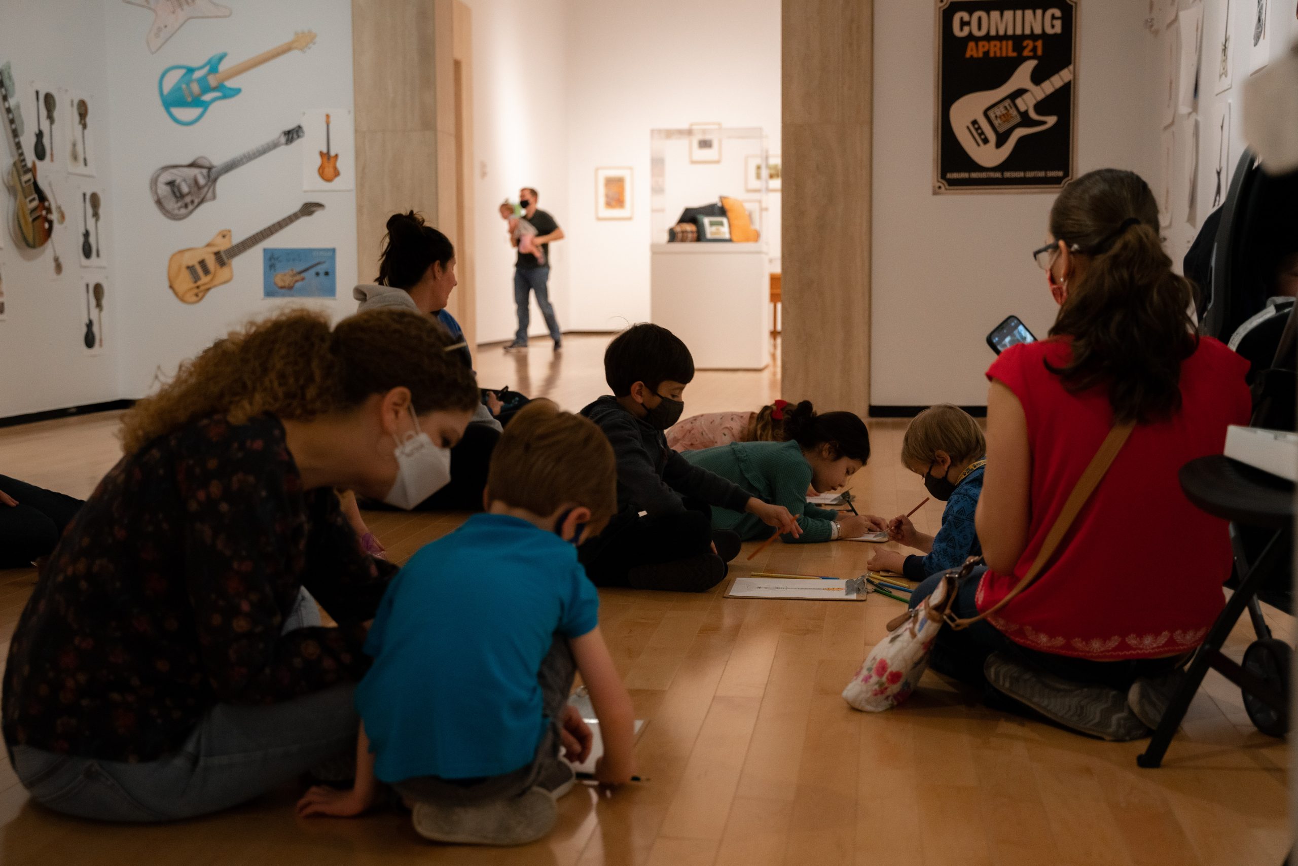 A group of children with their parents sit on the floor of a gallery while coloring on paper.