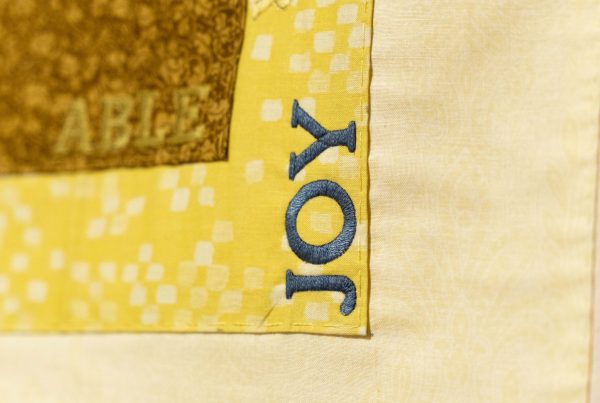 A square piece of fabric with the word joy stitched into it.