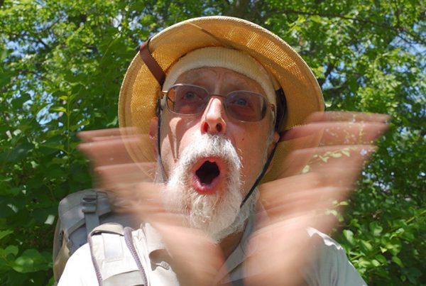 An elderly man in a safari outfit claps his hands and shouts.