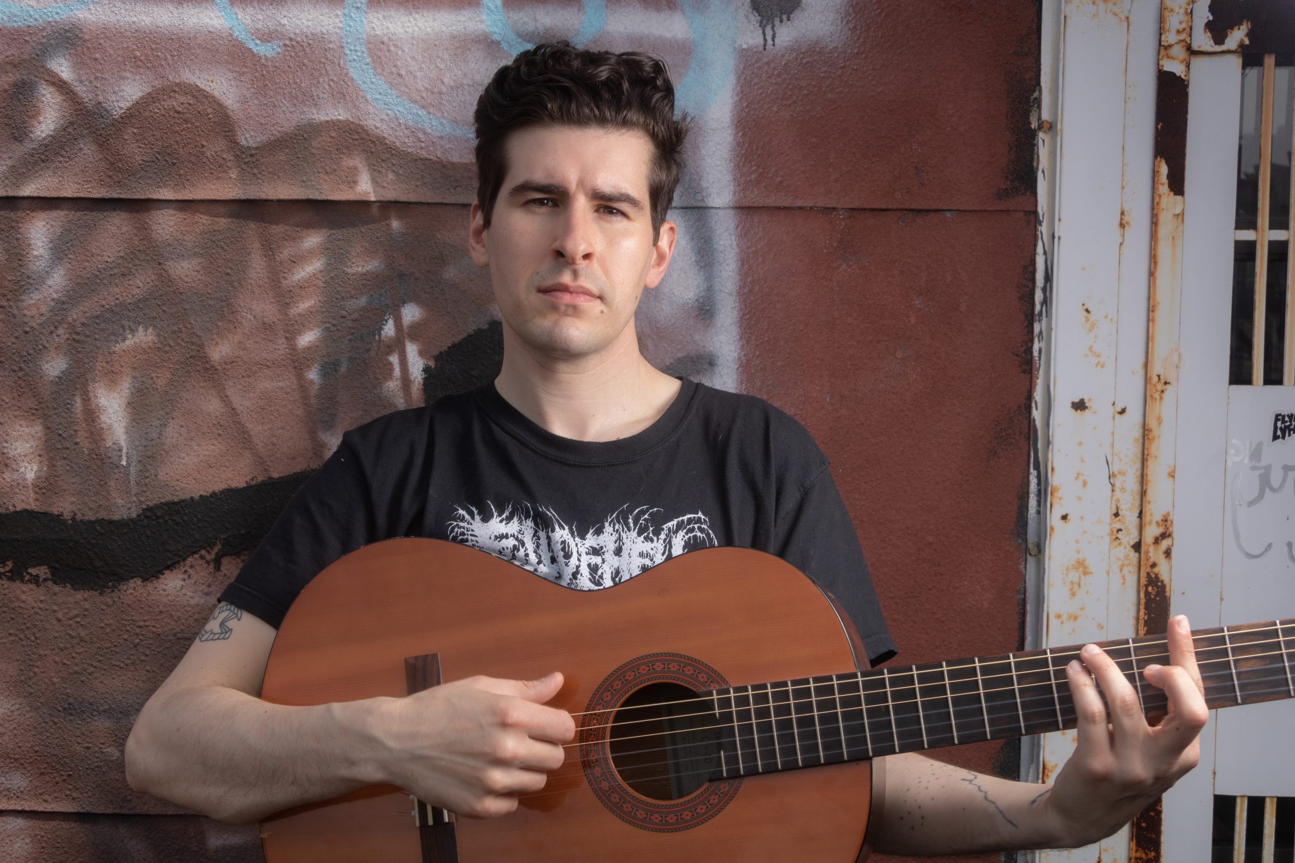 A man holds an acoustic guitar while leaning against a graffitied wall.