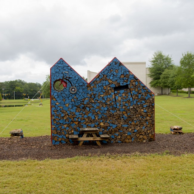 A house-like sculpture made of firewood, picnic table, desk, tricycle, paint, and steel pipe