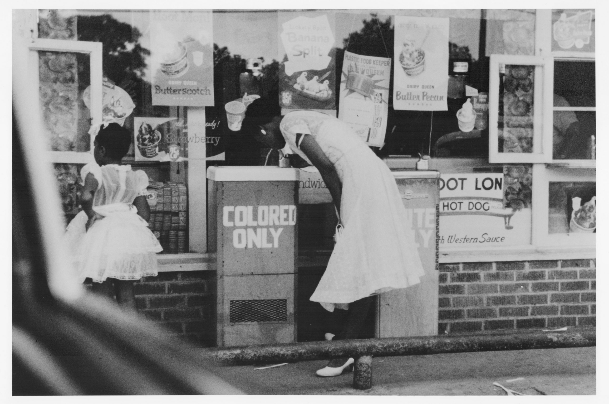 A woman takes a drink from a water fountain labeled "colored only."