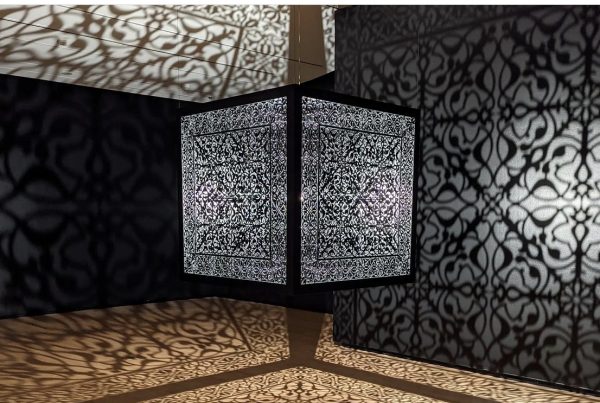 A large metal cube suspended from the ceiling, casting a cool complex pattern.