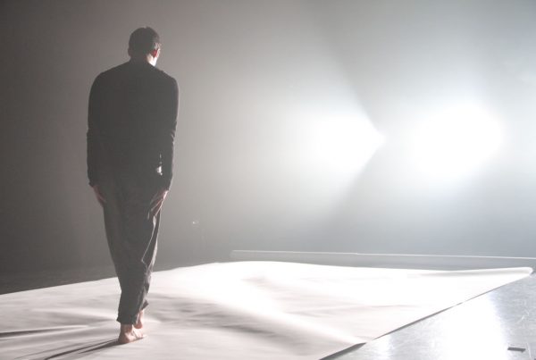 A figure with hands at their side looks down and walks toward bright lights.