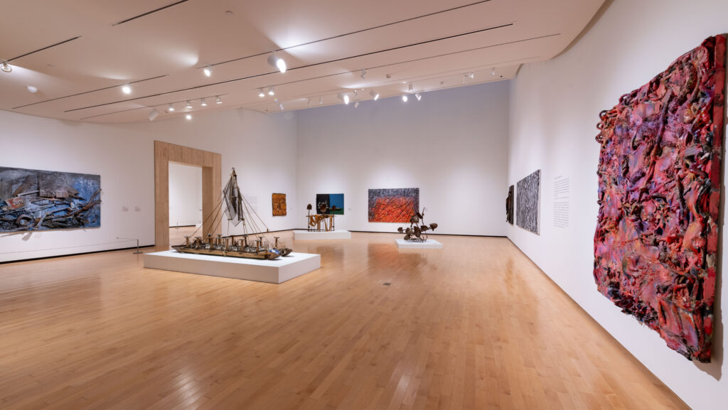 The Jule's exhibition, "Black Codes: Art and Post-Civil Rights Alabama," displayed in February 2024 in the Bill L. Harbert Gallery. Several mixed media works done on canvases hang on the walls with bright colors, and three sculptural works are in the center of the space, elevated on platforms. The sculptures are made with assortments of shovels, chainmail, and other found materials. One sculpture is made into a frigate-style ship.