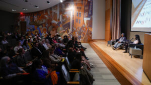 Audience members listen to exhibiting artist Lonnie Holley speak with guest curator Dr. Aleesa Pitchamarn Alexander at the inaugural Auburn Forum for Southern Art and Culture. Holley and Alexander are seated in chairs on stage while images of Holley's works are projected behind them, while audience members sit in auditorium seating, listening.