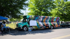The Jule Museum's Museum in Motion bus. Bus is turquoise colored overlaid with a series of arrows featuring details from artworks from The Jule's collection. Overtop the arrows is "Museum in Motion" in white lettering. Bus is parked alongside a street, featuring trees and people in a park.