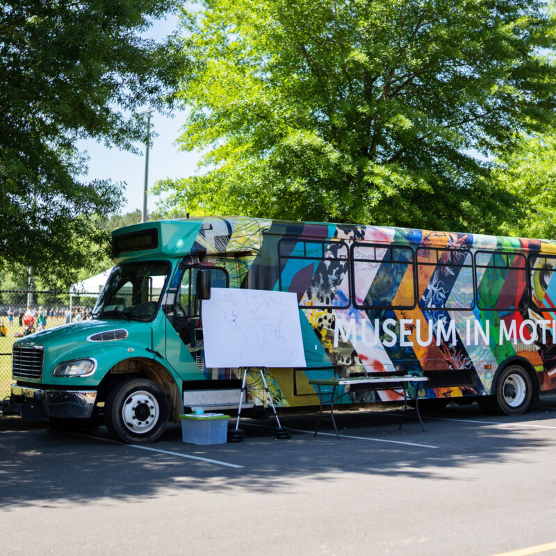 The Jule Museum's Museum in Motion bus. Bus is turquoise colored overlaid with a series of arrows featuring details from artworks from The Jule's collection. Overtop the arrows is "Museum in Motion" in white lettering. Bus is parked alongside a street, featuring trees and people in a park.