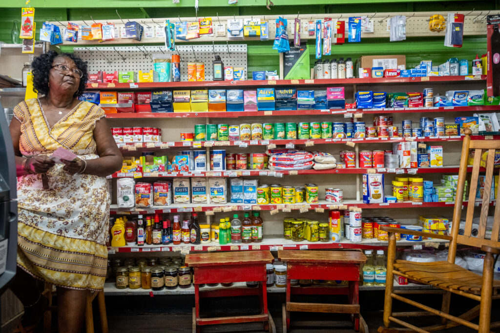 Photo of metal store shelves with a variety of goods for purchase ranging from canned vegetables to grits to over-the-counter medicine. A wooden rocking chair is featured on the right hand side, with a Black woman in a yellow, white, and brown floral patterned dress on the left, seated on a wooden stool. Photo by Kate Medley.