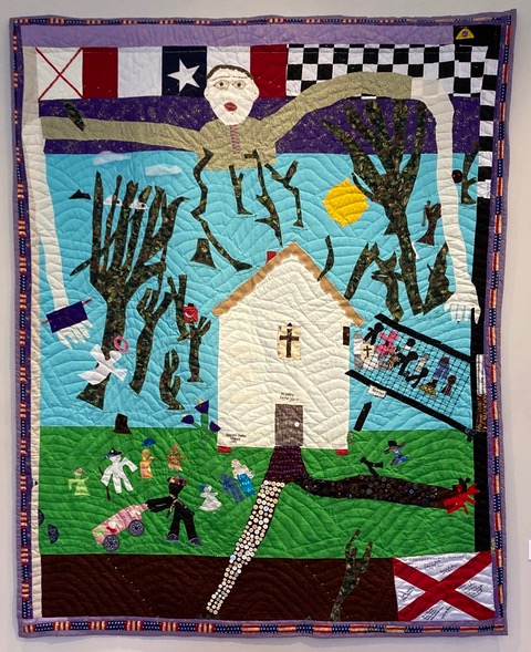 Quilt made by Yvonne Wells that depicts a scene featuring a white church building, black trees, small figures of people and a large, long-armed man in a tan shirt with brown hair reaching around the scene. Edges of the quilt featured American flag-style patterns, as well as a recreation of the Alabama state flag.