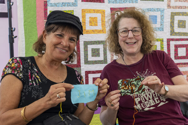 Two women smile and hold up their fabric with stitched names