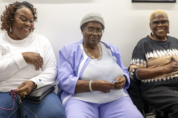 Three women smile as they stitch names into fabric.