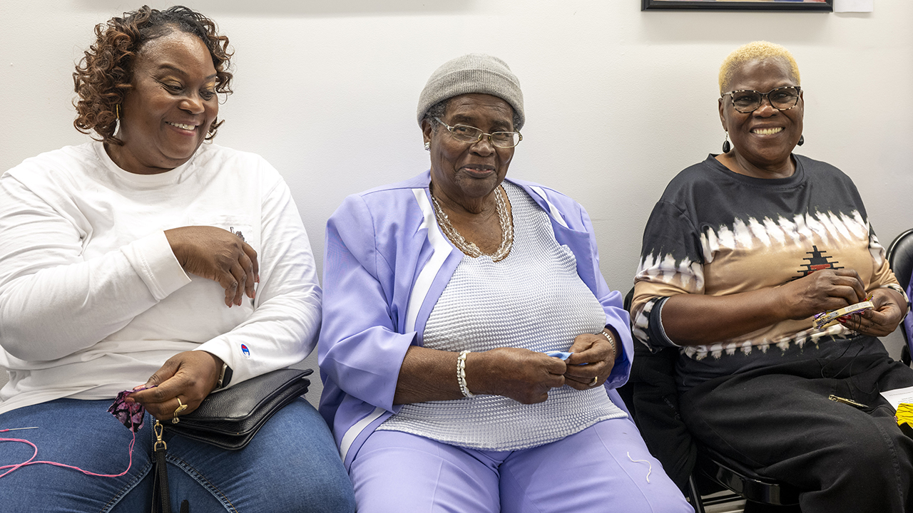 Three women smile as they stitch names into fabric.