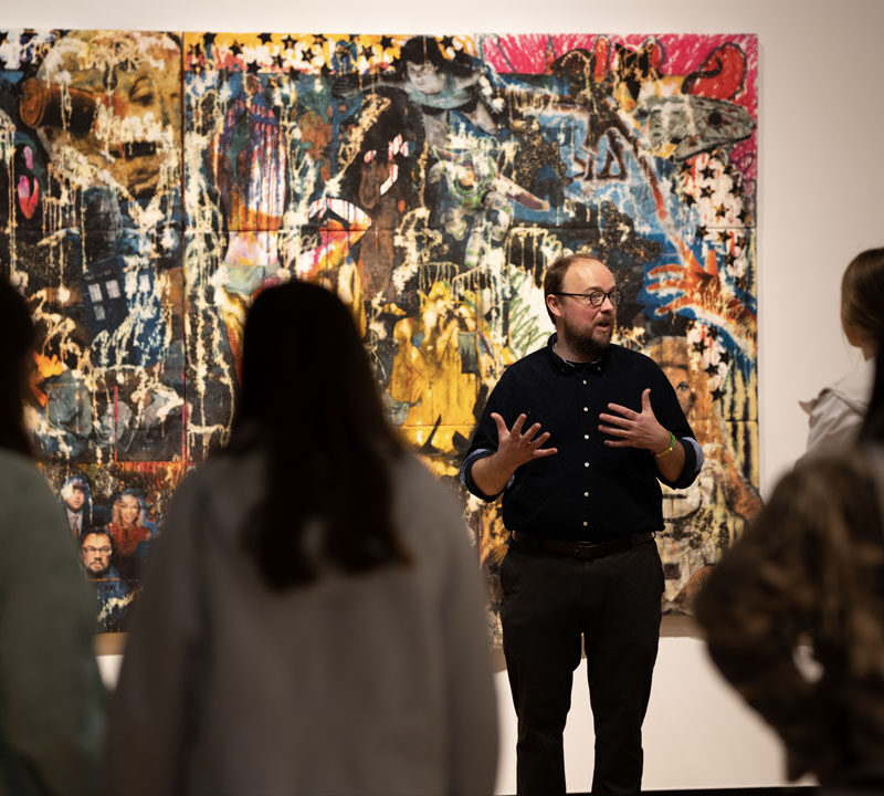 A person stands before a collage in a museum gallery, leading a tour of students.