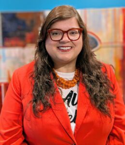 Headshot of Nancy Strickland Chavis. Strickland Chavis wears a bight read blazer, red framed glasses, and an orange beaded necklace, in front of a background of a painting in a gallery space.