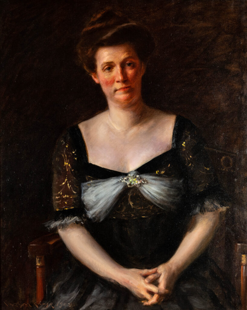A woman sits in a chair with her hands folded in her lap. She has a black dress with half sleeves and brown hair in an updo.
