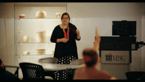Nancy Strickland Chavis teaches a museum studies course at UNC Pembroke. Strickland Chavis stands at the head of a classroom wearing a black shirt, a red beaded necklace, and a black and white polka-dotted skirt. A glass display cabinet behind her houses sculpted objects. In the foreground, a student raises her hand.