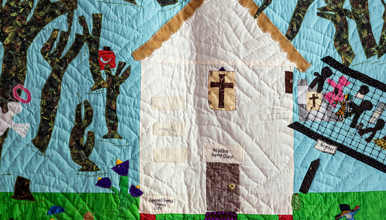 A vibrant quilt depicting a cream-colored church with a brown door and cross, set under a blue sky with yellow sun and dark trees. Green grass below shows colorful figures, a stroller, and a button-lined path leading to the house.
