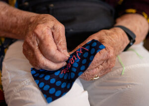A photo of hands sewing a name using red thread into a patch of black fabric with blue polka dots.
