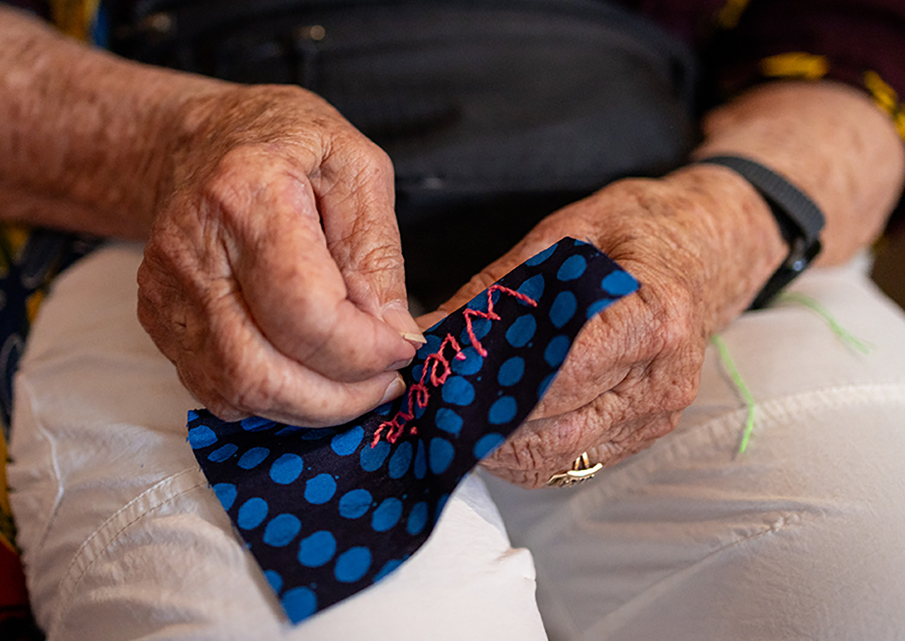 An older woman sews a name using red thread into a patch of black fabric with blue polka dots.