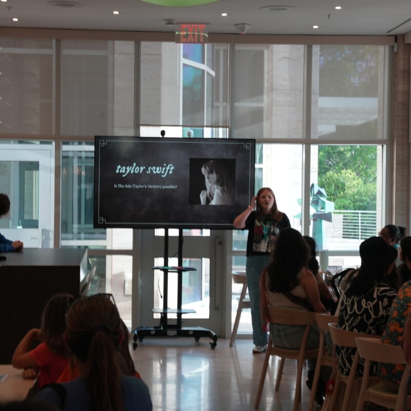 Eleanor Hickman at the beginning of her Taylor Swift-themed tour. She stands at the front of windows, speaking to people seated in a cafe space. Behind her, is a TV displaying a photo of Taylor Swift.
