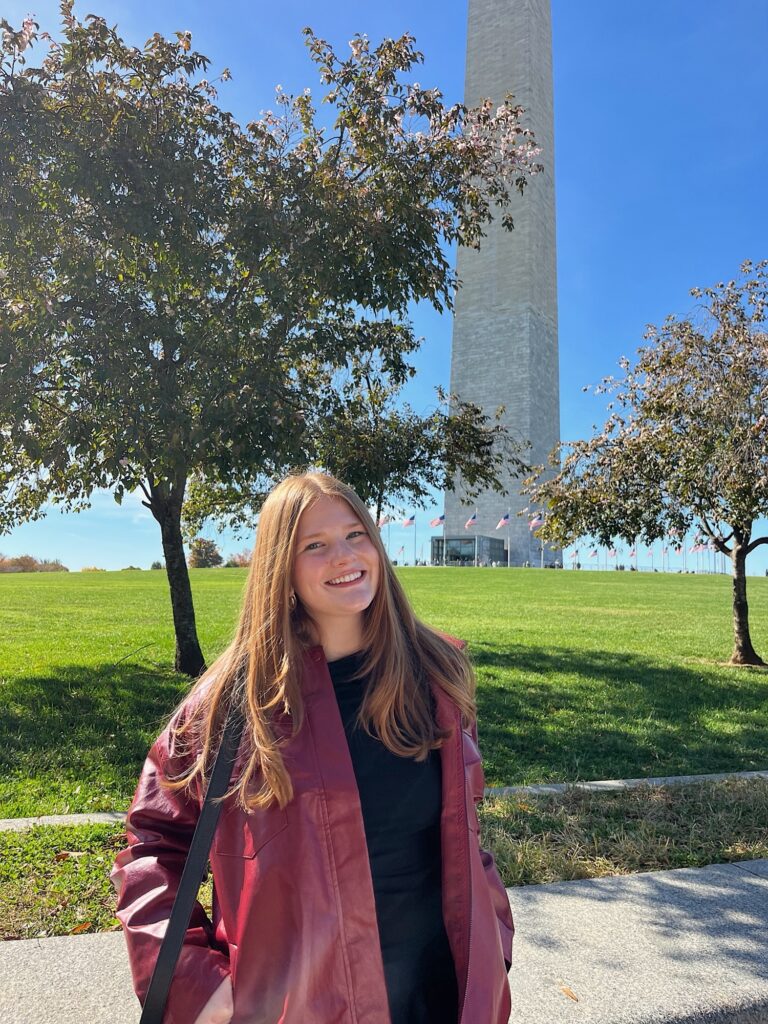 Photo of Eleanor Hickman standing in front of the Washington Memorial in Washington, D.C. She has long, strawberry blonde hair and wears a burgundy jacket.