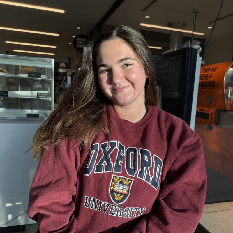 Comms & External Relations unit intern, Maggie Frost. Frost has shoulder-length brunette hair and wears a maroon crewneck sweatshirt with "Oxford University" and the crest embroidered in white and navy.