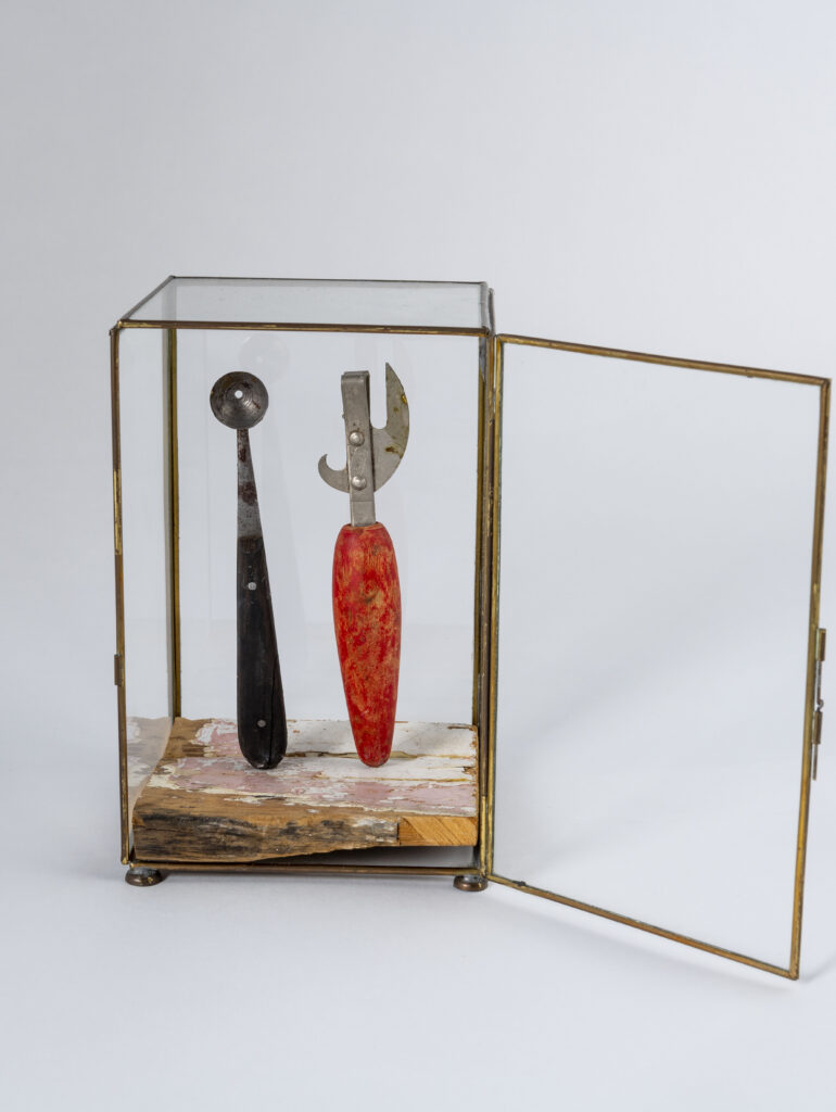 Two tools, a melon-baller with a black handle and an unknown tool with a red handle, set onto a piece of reused wood with remnants of white paint and placed inside a glass box with brass hinges which open to the front of the box.