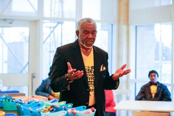 Rev. Dale Braxton opens the morning quilting workshops at the Auburn Forum. He wears a yellow t-shirt under a black blazer and stands in front of a table of quilting fabric and thread.