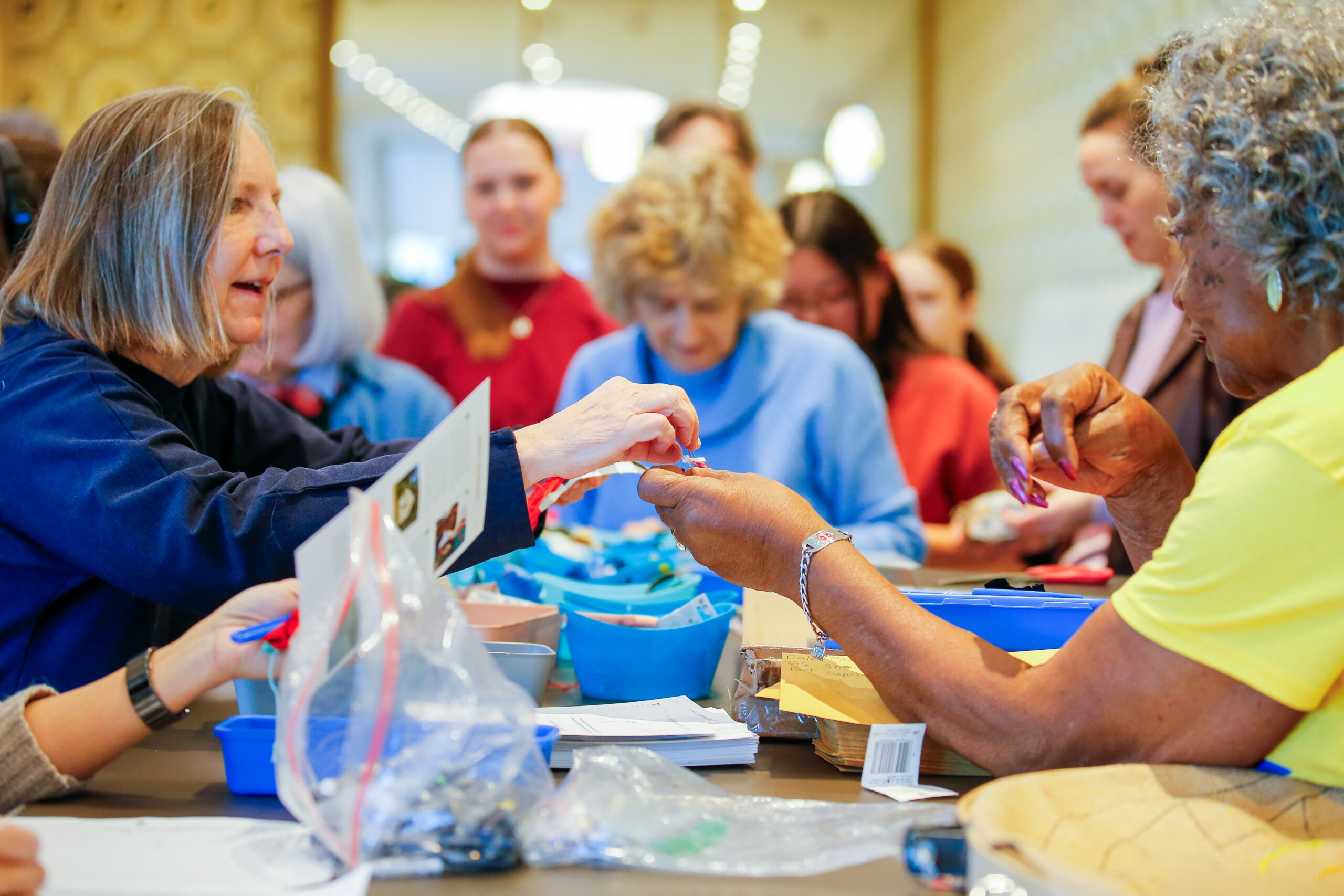 Auburn Forum participants work with the Mt. Willing Quilters. A woman in a yellow t-shirt holds out a needle to another woman in a long-sleeved blue shirt.
