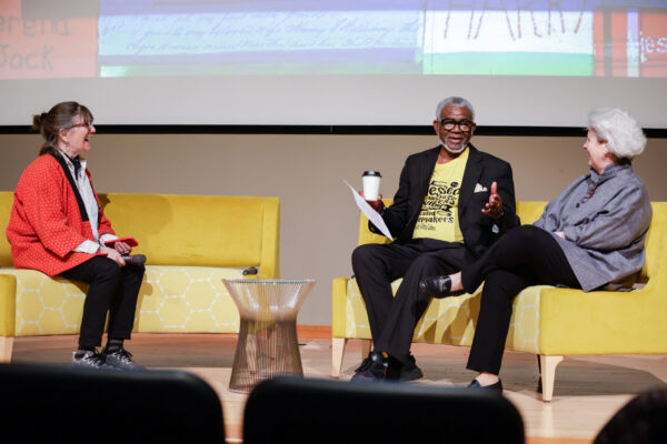 Forum moderator Nikki Silva (left) speaks with Rev. Dale Braxton (center) and Judge Susan Walker (right) about the origins of the Sew Their Names project. Silva wears a red jacket with black pants, Braxton wears black slacks and a black blazer with a yellow t-shirt underneath, and Walker wears a blue-grey blouse with black slacks.