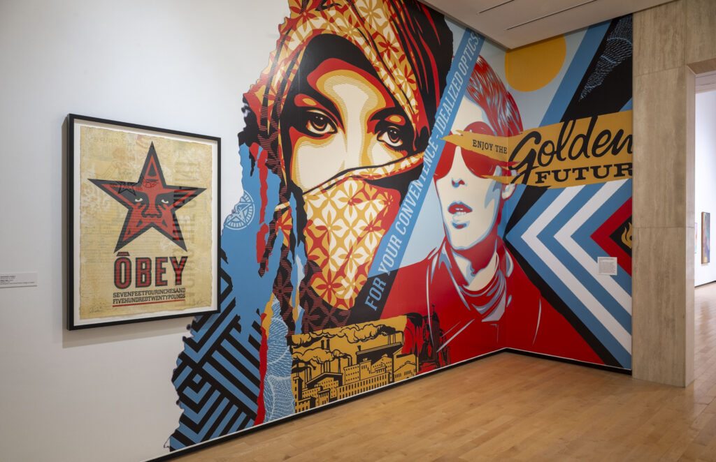Large-scale mural installation in a gallery showing stylized figures and bold geometric patterns in red, blue, black, and yellow. Visible text includes “For Your Convenient Personalized Optics” and “Enjoy the Golden Future,” alongside a framed OBEY poster featuring a red star.