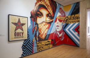 Large-scale mural installation in a gallery showing stylized figures and bold geometric patterns in red, blue, black, and yellow. Visible text includes “For Your Convenient Personalized Optics” and “Enjoy the Golden Future,” alongside a framed OBEY poster featuring a red star.