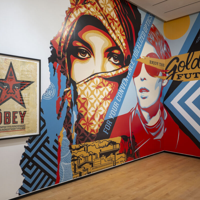 Large-scale mural installation in a gallery showing stylized figures and bold geometric patterns in red, blue, black, and yellow. Visible text includes “For Your Convenient Personalized Optics” and “Enjoy the Golden Future,” alongside a framed OBEY poster featuring a red star.