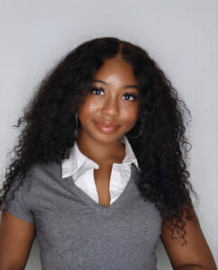 Photo of Kaitlin Stabler. Kaitlin is a young, Black woman with long loose curls, wearing a white collard shirt under a grey, short-sleeved sweater.